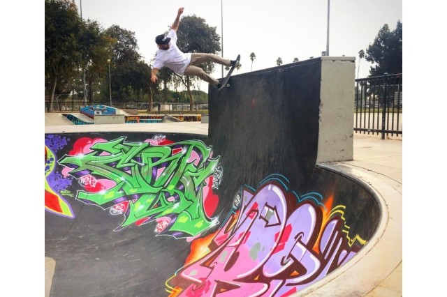 Man in white shirt and tan pants, skateboarding up an outdoor, concrete ramp covered in colorful graffiti.